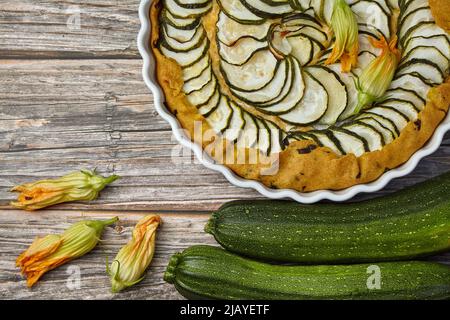Köstliche hausgemachte Zucchini und Käsekuchen umgeben von Blumen und Zucchini auf einem rauen Holztisch. Kochen Sie zu Hause Stockfoto