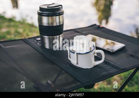 Trendy bequeme Papier Tropfen Kaffeetasche in Metallbecher auf Campingtisch im Freien. Frisch gebrühten Kaffee in der Natur zubereiten Stockfoto