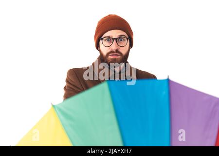 Oh, es regnet wieder. Europäischer bärtiger Kerl in einer Mütze schockiert hinter einem Regenbogenschirm. Wetterkonzept. Isoliertes Studio auf weißem Hintergrund. Hochwertige Fotos Stockfoto