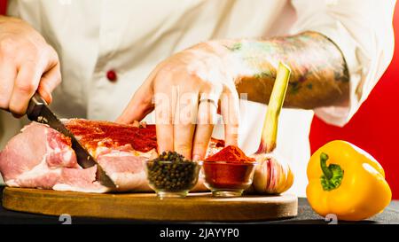 Großes Stück rohes Schweinefleisch auf Holzbrett mit Gewürzen, Gemüse. Nicht erkennbarer männlicher Koch schneidet Fleisch mit Messer. Nahaufnahme. Kochkonzept Stockfoto