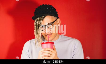 Junger hübscher stilvoller Mann in Gläsern, schwarze Kappe trinkt erfrischendes Getränk auf rotem Hintergrund. Hipster mit blonden Dreadlocks trinken Softdrink Th Stockfoto