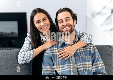 Porträt eines glücklichen kaukasischen Paares, Männer und Frauen, die Zeit miteinander verbringen, sich zu Hause auf der Couch im Wohnzimmer sitzend umarmen, auf die Kamera schauen, glücklich lächeln, harmonisches Beziehungskonzept Stockfoto