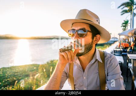 Lateinischer Hipster in einer Bar am Ufer eines Sees bei Sonnenuntergang mit Hut und Hosenträger und Zigarre rauchendem Rauchen Stockfoto