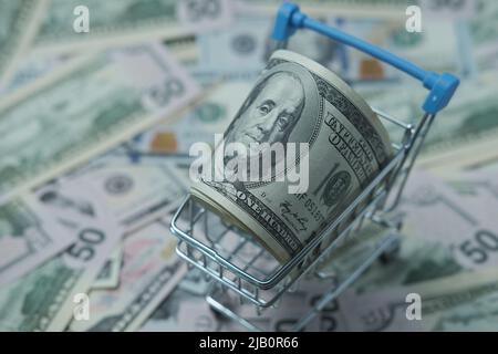 In einen Schlauch gerollte Dollars liegen in einem Einkaufskorb. Der Korb steht auf einem verschwommenen Geldhintergrund. Stockfoto