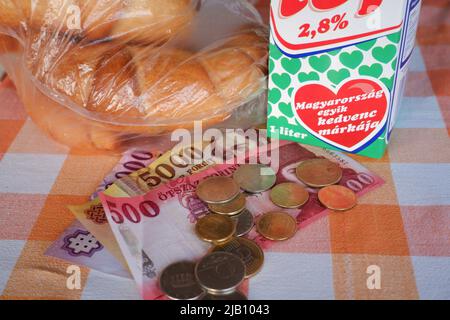 Budapest, Ungarn - 26. Mai 2022: Grundnahrung (Milch, Croissant), ungarische Forint-Banknoten und -Münzen auf einem rot-weißen karierten Tisch in einem Haus Stockfoto