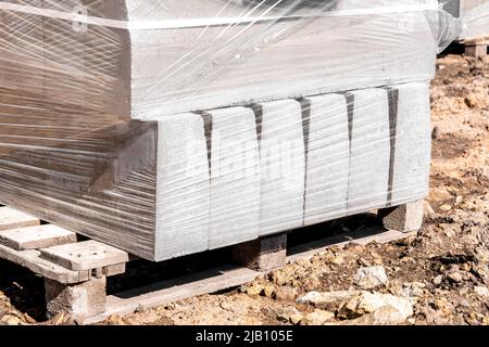 Ein Bordstein in Transportverpackungen steht auf einer Holzpalette auf dem Lehmboden einer Baustelle, selektiver Fokus Stockfoto