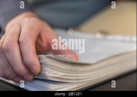 Eine Nahaufnahme der Hand eines Mannes, die in einem Ordner durch Dokumente blättert. Ein erwachsener Mann hält einen Bericht in einem geöffneten grauen Ordner mit einem Ordner. Selektives F Stockfoto