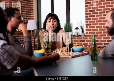 Glückliche Frau, die am Tisch sitzt und Schach spielt, während sie im Wohnzimmer auf die Kamera schaut. Verschiedene Menschen sitzen zu Hause und entspannen sich bei Strategiespielen, während sie Getränke und Snacks genießen. Stockfoto