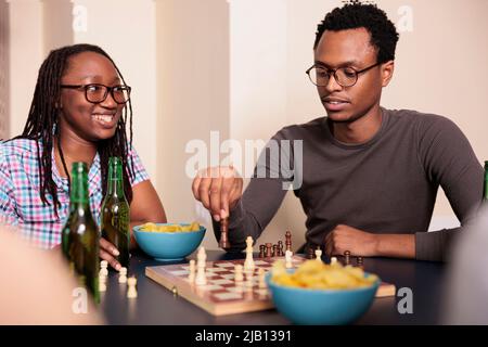 Lächelnde Frau, die einen nachdenklichen Freund ansieht, der das Schachstück an Bord bewegt. Zwanglose Menschen entspannen sich bei Strategiespielen, während sie gemeinsam zu Hause im Wohnzimmer Spaß haben. Stockfoto