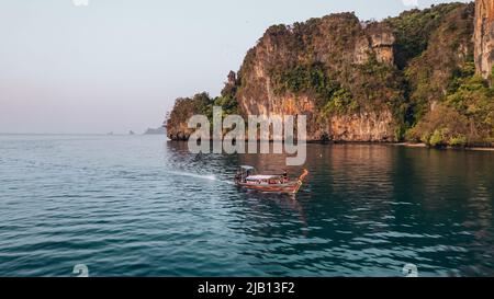 Sonnenaufgang am Railay Beach in Krabi Thailand mit einem thailändischen Longtail-Boot, das an Kalksteinfelsen im Andamanensee vorbeifährt Stockfoto