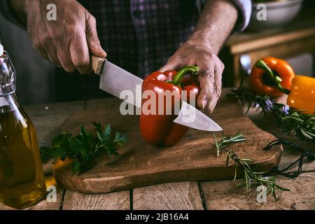 Hoher Winkel der Ernte gesichtslosen männlichen Chef hacken frischen reifen roten Paprika mit Messer auf Holz Schneidebrett weiß Vorbereitung gesunder Salat in der Küche Stockfoto