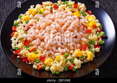 Gekochte kleine rosafarbene Garnelen auf Hirse-Pilaf mit Gemüse, Mangostückchen und Gewürzen auf schwarzem Teller auf dunklem Tisch, horizontale Ansicht von oben, Stockfoto