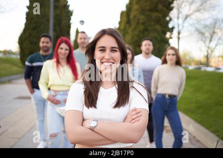 Selbstbewusste Frau, die die Kamera anschaut und lächelt, während sie mit gekreuzten Armen vor einer Gruppe von Menschen steht. Freundschafts-, Union- und Teamkonzept. Stockfoto