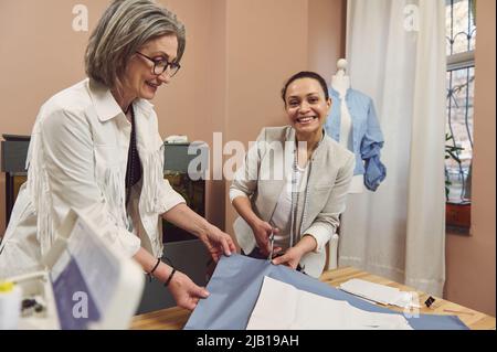 Taillenlang Porträt von lächelnden, kreativen, qualifizierten, inspirierten reifen weiblichen Modedesignerinnen Schneidern Herstellung Kleidung Muster auf Stoff, während Nähen c Stockfoto