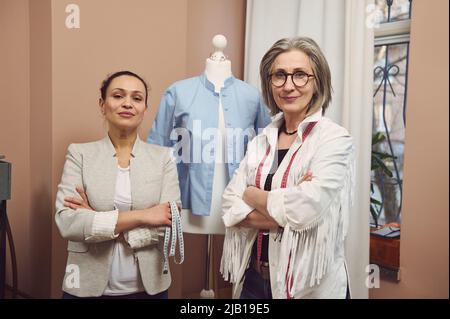 Ein erfahrenes, inspiriertes Team aus zwei Näherinnen unterschiedlichen Alters und Nationalitäten, die mit gekreuzten Armen mit einer Schaufensterpuppe mit blauem Shi posieren Stockfoto