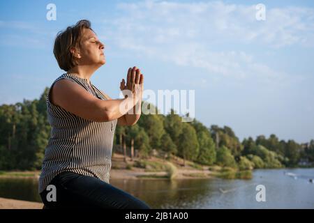 Einsame Frau meditiert am Ufer des Sees mit ihren Handflächen, die vor ihr zusammengekrallt sind, oder betet. Das Konzept der Entspannung und des spirituellen Gleichgewichts und der Verbindung mit der Natur Stockfoto