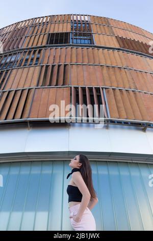 Blick von unten selbstbewusste junge Frau in modischem Outfit mit Blick auf die Kamera, während sie in der Nähe eines modernen Gebäudes in der Stadt steht Stockfoto