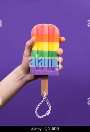 Die Hand des jungen Mannes hält einen regenbogenfarbenen Schaumschwamm auf einem violetten Hintergrund. Stockfoto
