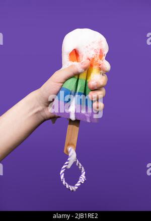 Die Hand des jungen Mannes hält einen regenbogenfarbenen Schaumschwamm auf einem violetten Hintergrund. Stockfoto
