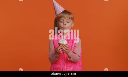 Happy Kleinkind Kinder Mädchen tragen festliche Kegelkappe Geburtstag Feier Party, weht Kerze auf kleinen Kuchen Cupcake einen Wunsch. Kleines Kind auf orangefarbenem Hintergrund isoliert Stockfoto