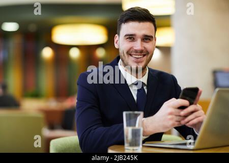 Positiv aufgeregt jungen Geschäftsmann mit Stoppeln sitzt am Tisch im Restaurant und arbeitet an Online-Projekt, er mit Smartphone und Laptop Stockfoto
