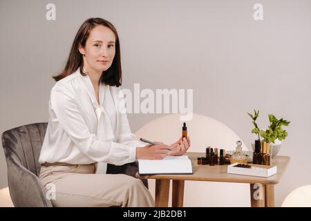 Das Aromatherapeutin-Mädchen sitzt in ihrem Büro und hält eine Flasche aromatisches Öl in den Händen und schreibt etwas auf. Es gibt ätherisches Öl Stockfoto