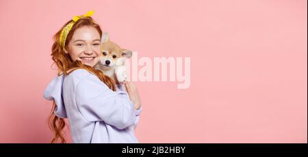 Fröhliche Rothaarige junge Mädchen mit langen lockigen Haaren hält niedlichen kleinen Welpen von Corgi Hund isoliert auf rosa Hintergrund. Konzept von Jugend, Schönheit Stockfoto