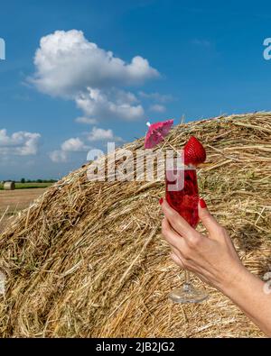 Eine weibliche Hand mit einem Kelch, der einen Erdbeercocktail enthält, gegen einen Heuballen unter einem wunderschönen Himmel Stockfoto