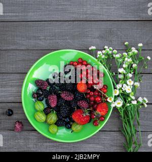 Beerenmischung - Johannisbeeren, Stachelbeeren, Maulbeeren, Erdbeeren auf einem Teller neben einem Strauß wilder Blumen auf einem hölzernen Hintergrund mit Kopierfläche. Sommer Stockfoto