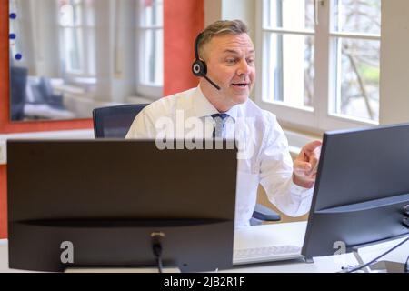 Ein Geschäftsmann, der ein Headset in einer Telefonkonferenz trägt, redet und gestikelt auf seinem Computerbildschirm in einem Büro Stockfoto