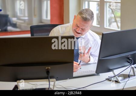 Frustrierter Geschäftsmann, der in einem Büro zwischen Computermonitoren ein Wringen mit den Händen ansieht Stockfoto