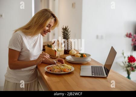 Blonde hübsche Frau, die Pasta mit Garnelen in der Küche am Laptop isst. Ein Glas Wein zum Mittagessen. Stockfoto