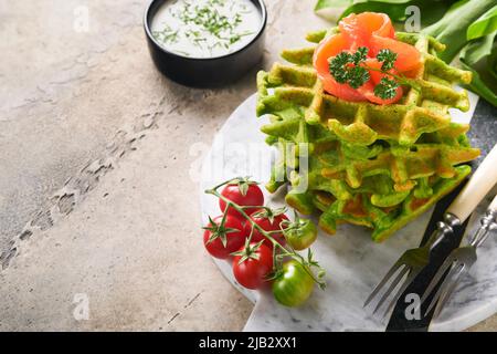 Grüne belgische Waffeln. Spinat- oder Bärlauch- oder Pesto-Waffeln mit rotem Lachs und Sahnesauce auf grauem Beton-Tischhintergrund. Köstliches Frühstück, Stockfoto