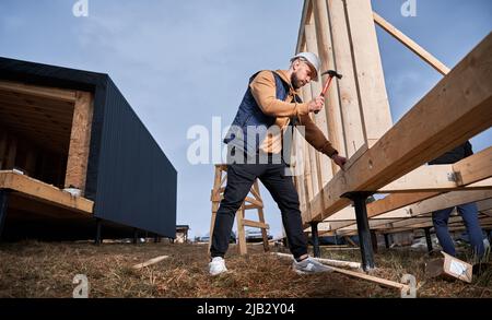 Mann Arbeiter Gebäude Holzrahmenhaus auf Pfahlfundament. Zimmermann hämmert mit Hammer Nagel in Holzbrett. Schreinerei-Konzept. Stockfoto