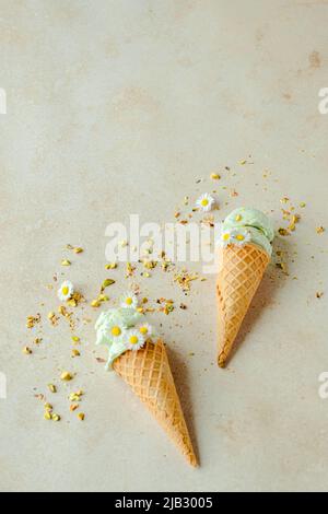 Pistazien-Eis in einem Horn ist mit Kamillenblüten und verstreuten Nüssen verziert. Sommer saisonal kalt süß gesunde Kinder Dessert. Flach Lay Stockfoto