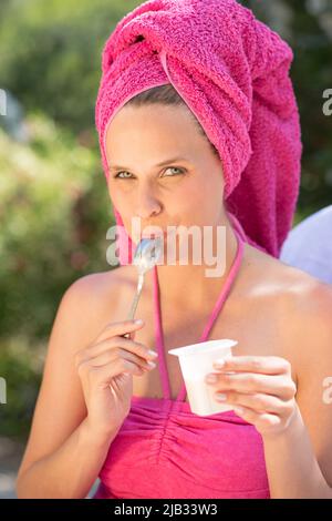 Junge Frau in einem rosa Badeanzug sitzt mit einem Handtuch am Kopf draußen und genießt einen frischen Joghurt Stockfoto