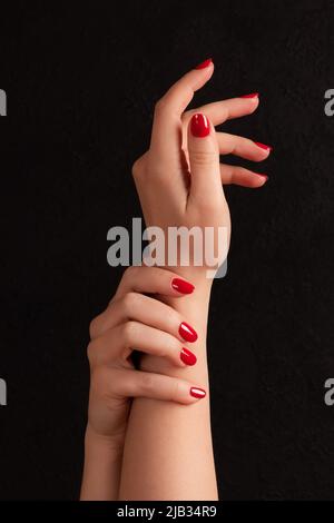 Weibliche Hände mit schöner Maniküre - rote Nägel auf dunkelschwarzem Hintergrund. Nagel, Hautpflegekonzept Stockfoto