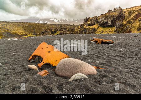 Zwei verrostete Wrackfragmente liegen im schwarzen Sand des Djúpalónssandur zwischen gerundeten Kieselsteinen, Halbinsel Snæfellsnes, Island, mit moosbedeckten Lavaklippen und schneebedeckten Bergen im Hintergrund. Das Wrack gehört zum britischen Kutter Epine GY 7, der in der Nacht des 13. März 1948 östlich von Dritvík auflief. Vierzehn der neunzehn Besatzungsmitglieder starben, fünf wurden gerettet. Die Überreste sind im Snæfellsjökull-Nationalpark geschützt. Stockfoto