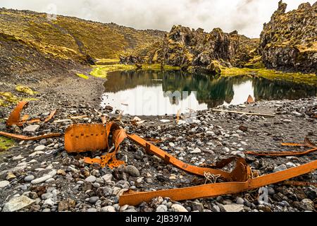 Am Rand eines kleinen, ruhigen Teiches am Djúpalónssandur, Halbinsel Snæfellsnes, Island, spiegeln sich die moosbedeckten Lavaklippen im Wasser, liegen verrostete Rippen und Streben. Das Wrack gehört dem britischen Traupler Epine GY 7, der in der Nacht des 13. März 1948 östlich von Dritvík strandete. Vierzehn der neunzehn Besatzungsmitglieder starben, fünf wurden gerettet. Die Überreste sind im Snæfellsjökull-Nationalpark geschützt. Stockfoto