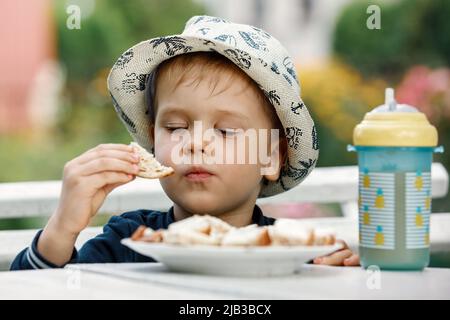 Ein gesundes Kind isst in den Sommerferien im Garten seiner Großmutter Butter auf Brot. Ein Plastikbecher mit Ananassaft auf dem Tisch. Die Konz Stockfoto