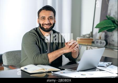 Porträt eines gutaussehenden, erfolgreichen arabischen oder indischen Mannes in lässiger, stilvoller Kleidung, Finanzexperte, Aktienhändler, der an seinem Schreibtisch sitzt, die Kamera anschaut und freundlich lächelt Stockfoto