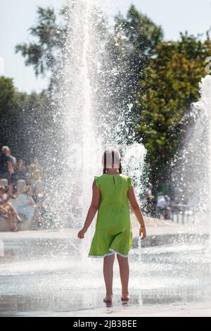 Barfuß Mädchen in grünem Kleid vor einem Brunnen im Park. Hohe Wasserströmung, Spaß für Kinder an einem heißen Sommertag. Stockfoto