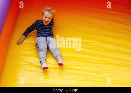 Ein urkomischer kleiner Junge rutscht mit hoher Geschwindigkeit von einem großen, leuchtend gelben Trampolin herunter. Kind, das unterwegs ist, steht auf dem Foto Platz für Text zur Verfügung Stockfoto