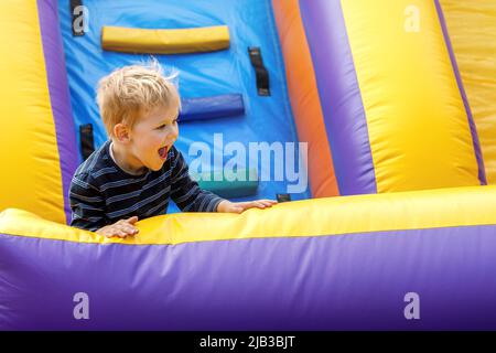 Fröhliches Kind, das auf einem farbenfrohen aufblasbaren Trampolin springt. Kleiner Junge, 3-4 Jahre, der an sonnigen Sommertagen auf einem Spielplatz im Freien spielt. Kinder haben Spaß bei Aktivitäten Stockfoto
