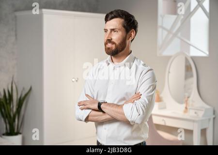 Seitenansicht des Brünetten Verkäufers, Designers, Architekten, der mit gekreuzten Händen drinnen steht. Schöner Mann mit lächelndem Bart, der im Schlafzimmer nach vorne blickt. Konzept der Gestaltung. Stockfoto