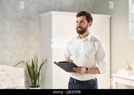 Seitenansicht von brünett männlich mit Bart Gestaltung Möbel, Interieur. Schöner Verkäufer, Designer, Architekt stehend, Ordner halten, schreiben, Suchen nach oben. Konzept der Gestaltung. Stockfoto