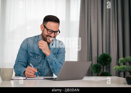 Glücklicher Unternehmer, der erfolgreichen Geschäftsplan in Tagebuch schreibt. Der Profi arbeitet am Schreibtisch am Laptop. Im Büro trägt er elegante Casuals. Stockfoto