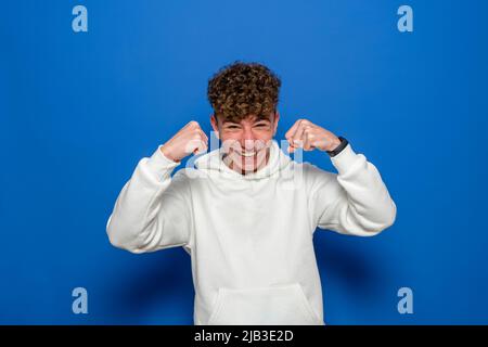 Starke sportlich schockiert jungen Mann 20s trägt weißes Sweatshirt stehend zeigt Bizeps Muskeln schaut auf Kamera isoliert auf blauem Hintergrund Studio Stockfoto