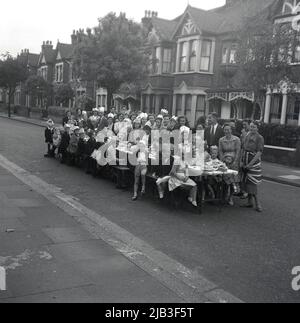 1953, eine historische Straßenparty, eine Gruppe von Männern, Frauen und Kindern versammeln sich um einen langen Tisch auf der Straße für ihr Foto, während sie die Krönung von Königin Elizabeth II. Auf den Thron, England, Großbritannien, feiern. Da in der Nachkriegszeit nur wenige einen Fernseher hatten, gingen Menschen und Gemeinden auf die Straße, um zu feiern, eine Gelegenheit für Menschen jeden Alters, sich zusammenzufinden. Stockfoto