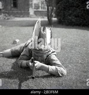 1950s, historisch, draußen im Garten, ein Junge, der spielt, vorgibt, ein gebürtiger oder roter inder zu sein, auf dem Gras liegend, mit seinem Spielzeuggewehr, Federhut und gemaltem Gesicht, England, Großbritannien. In Großbritannien nach dem Krieg war das Spielen von Cowboys und indianern ein beliebtes Spiel für junge Kinder. Stockfoto